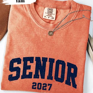 Pode incluir: Uma t-shirt de manga curta cor de inhame com decote redondo. A pe&ccedil;a apresenta a palavra SENIOR em letras mai&uacute;sculas grandes de azul-marinho desgastado, com o n&uacute;mero 2027 impresso por baixo numa fonte azul-marinho a condizer.