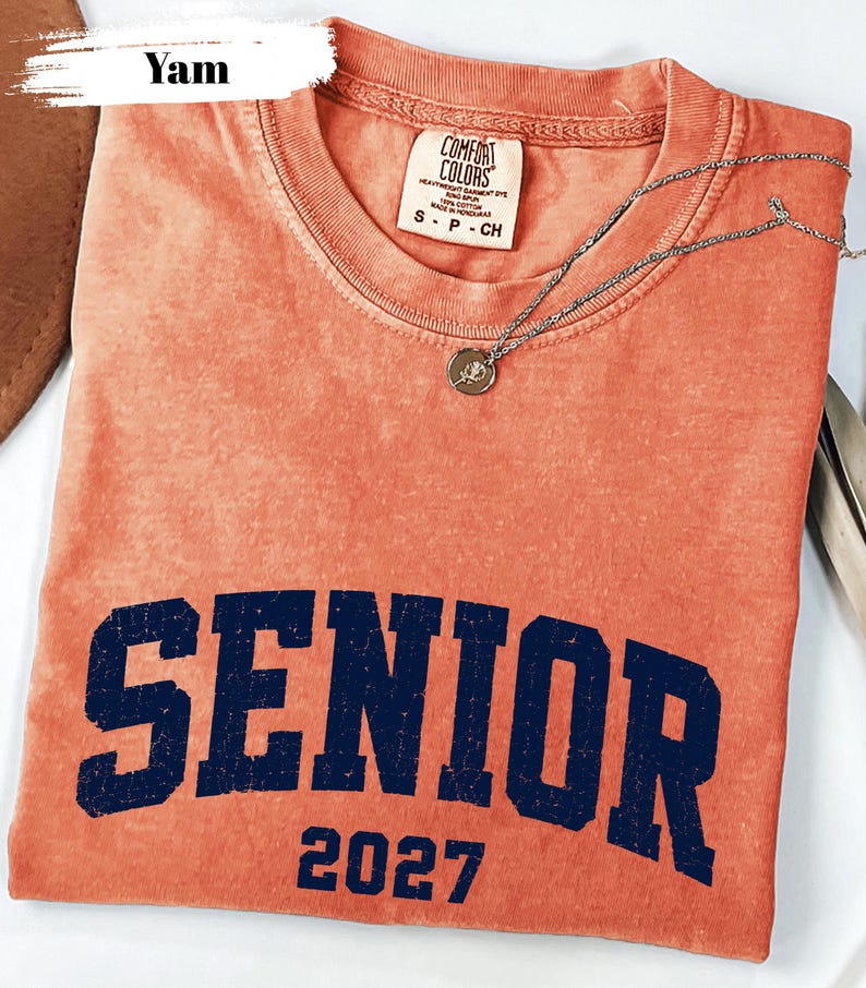 Pode incluir: Uma t-shirt de manga curta cor de inhame com decote redondo. A pe&ccedil;a apresenta a palavra SENIOR em letras mai&uacute;sculas grandes de azul-marinho desgastado, com o n&uacute;mero 2027 impresso por baixo numa fonte azul-marinho a condizer.