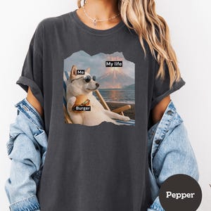 May include: Dark gray t-shirt featuring a graphic of a dog wearing sunglasses, holding a burger, and relaxing on a beach chair. The graphic includes the words "Me", "Burger", and "My life". The shirt is labeled "Pepper".