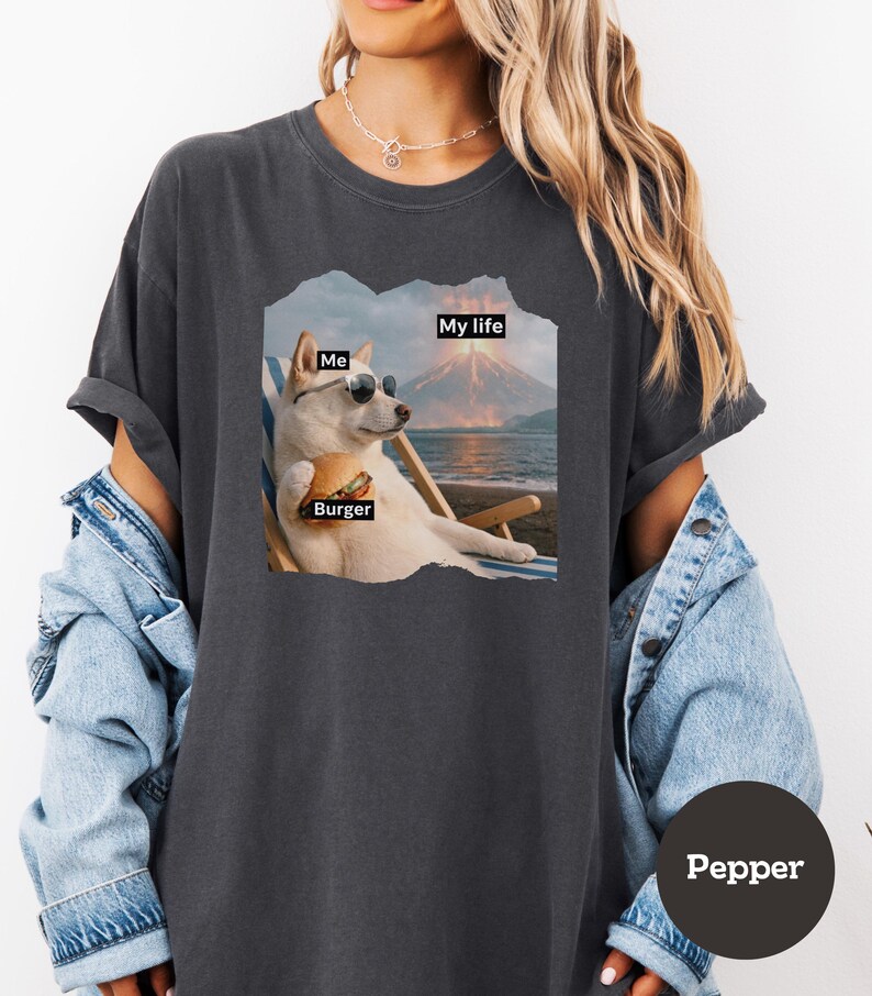 May include: Dark gray t-shirt featuring a graphic of a dog wearing sunglasses, holding a burger, and relaxing on a beach chair. The graphic includes the words "Me", "Burger", and "My life". The shirt is labeled "Pepper".