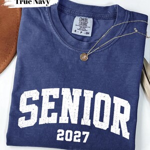 Pode incluir: Uma t-shirt de manga curta azul-marinho com um gr&aacute;fico branco desgastado que diz SENIOR 2027. A pe&ccedil;a apresenta gola redonda e uma etiqueta da marca Comfort Colors no interior. Um colar de corrente dourada fina com um pequeno pendente redondo repousa sobre o tecido.