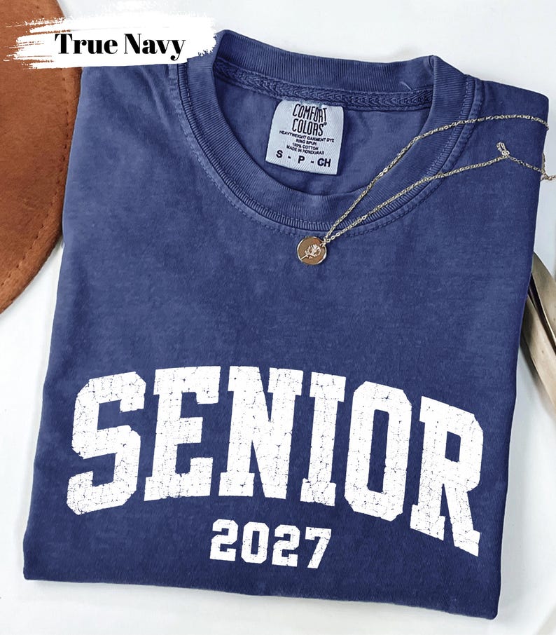 Pode incluir: Uma t-shirt de manga curta azul-marinho com um gr&aacute;fico branco desgastado que diz SENIOR 2027. A pe&ccedil;a apresenta gola redonda e uma etiqueta da marca Comfort Colors no interior. Um colar de corrente dourada fina com um pequeno pendente redondo repousa sobre o tecido.