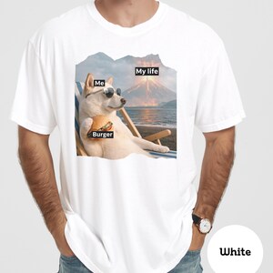 May include: White t-shirt featuring a dog wearing sunglasses, holding a burger, and relaxing in a beach chair. The shirt has the text "Me", "Burger", and "My life" in black boxes, with a volcano and ocean background.