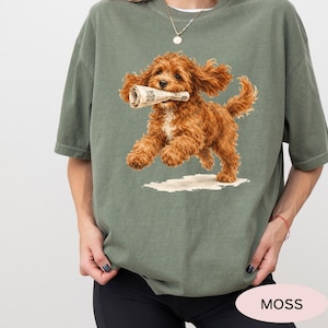 May include: A moss green short-sleeve t-shirt featuring a graphic of a fluffy brown dog running while holding a rolled-up newspaper in its mouth. The dog has curly fur and is depicted in a dynamic pose against a plain background.