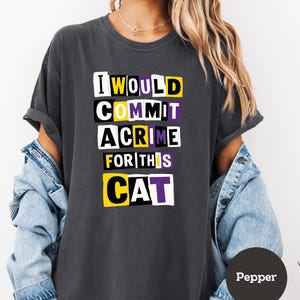 May include: Dark gray t-shirt with the text "I WOULD COMMIT A CRIME FOR THIS CAT" in a bold, block letter design. The text features yellow, purple, and white accents. The shirt is a casual style.