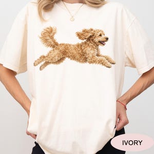 May include: An ivory-colored short-sleeve t-shirt featuring a graphic illustration of a golden-brown curly-haired dog in a running pose. The dog is depicted with its mouth slightly open and ears flowing back as if in motion.
