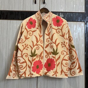May include: A cream-coloured jacket featuring vibrant embroidery of large pink flowers with green centres and swirling brown vine patterns. The garment has a high collar, long sleeves, and an open front design with visible striped lining on the interior.