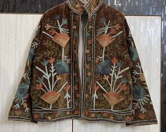 Op de afbeelding: Een bruin fluwelen jasje met een ingewikkeld geborduurd bloemenpatroon in tinten oranje, blauwgroen en lichtgroen. Het jasje heeft een hoge kraag, lange mouwen en een verticaal gestreepte voering die zichtbaar is bij de opening aan de voorzijde.