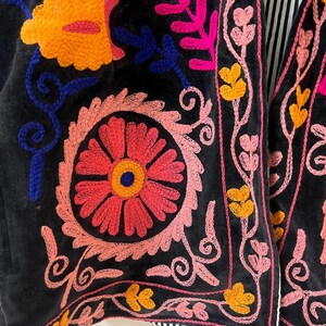以下が含まれることがあります： 鮮やかな花の刺繍が施された黒いベストのクローズアップ。刺繍はピンク、オレンジ、黄色、青の糸を使用し、カラフルで複雑なデザインを作り出しています。ベストにはストライプの裏地が付いています。