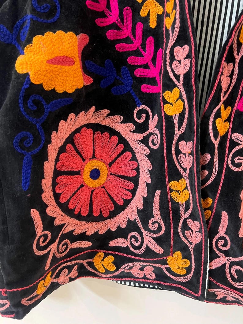以下が含まれることがあります： 鮮やかな花の刺繍が施された黒いベストのクローズアップ。刺繍はピンク、オレンジ、黄色、青の糸を使用し、カラフルで複雑なデザインを作り出しています。ベストにはストライプの裏地が付いています。