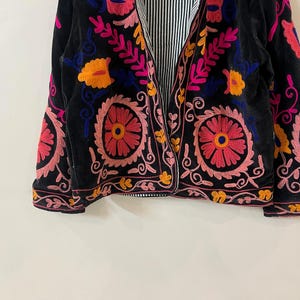 以下が含まれることがあります： ピンク、オレンジ、青色の鮮やかな花の刺繍が施された黒いベルベットのジャケット。 ジャケットの内側の裏地は、黒と白のストライプ模様です。 刺繍のデザインには、円形と花のモチーフが含まれており、ボヘミアンな美学を作り出しています。