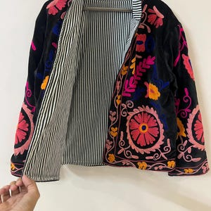 以下が含まれることがあります： ピンク、オレンジ、青色のカラフルな花の刺繍が施された黒いベルベットのジャケット。 ジャケットは、黒と白のストライプの裏地とスタンドカラーが特徴です。 刺繍は詳細で、前面と袖を覆っています。