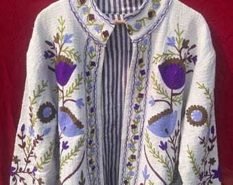 May include: A light-colored jacket with floral embroidery in purple, blue, and brown. The jacket has a striped lining and is displayed on a wooden hanger. The embroidery features stylized flowers and leaves.