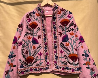 May include: A pink embroidered jacket with a floral design. The jacket features a stand-up collar and long sleeves. The embroidery includes red, orange, blue, and purple flowers and leaves. The jacket is hanging on a wooden hanger.