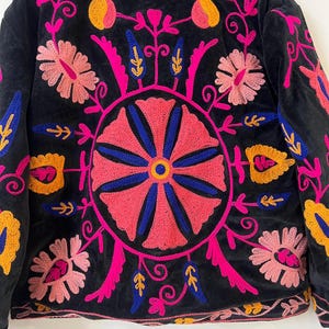 以下が含まれることがあります： ピンク、青、黄色で鮮やかな花の刺繍が施された黒いジャケット。ジャケットは背中に丸い花の模様があり、襟と袖には複雑な模様があります。刺繍は細かく、カラフルで、印象的な視覚的コントラストを生み出しています。