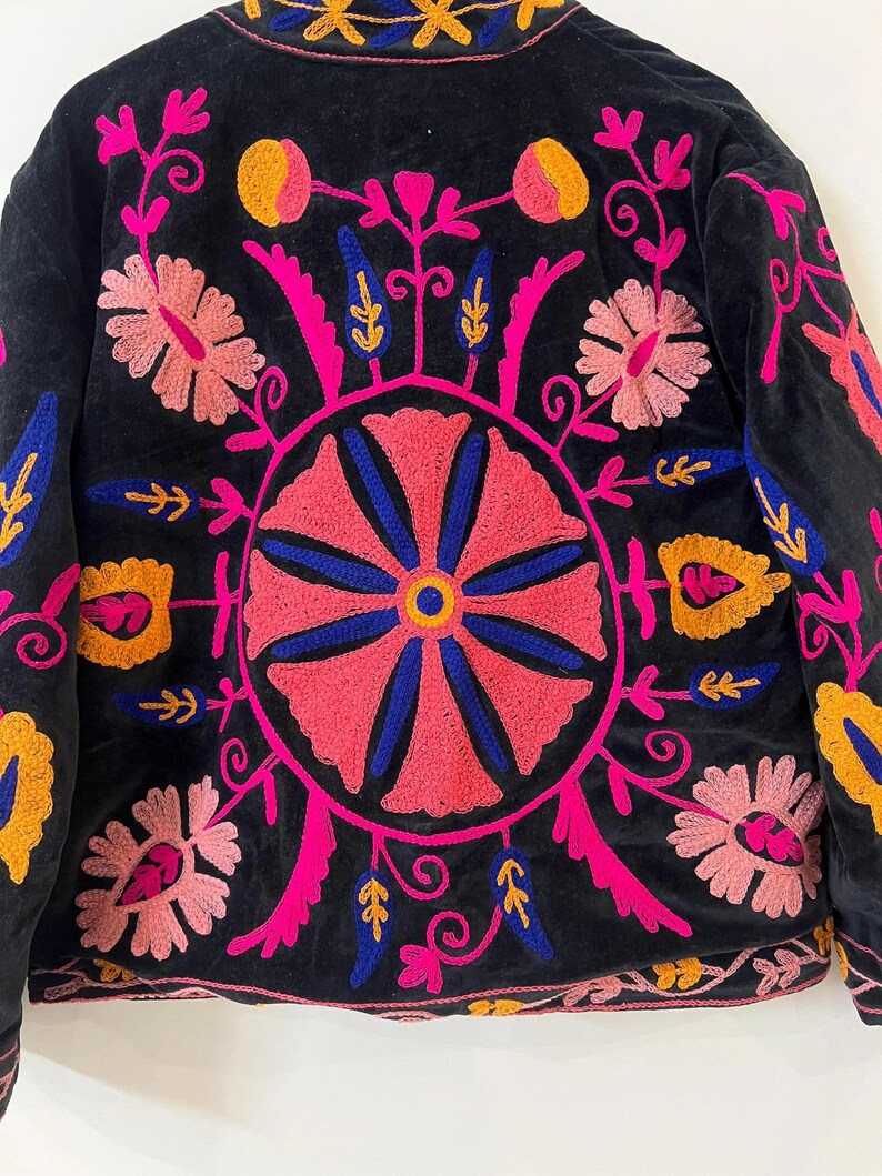 以下が含まれることがあります： ピンク、青、黄色で鮮やかな花の刺繍が施された黒いジャケット。ジャケットは背中に丸い花の模様があり、襟と袖には複雑な模様があります。刺繍は細かく、カラフルで、印象的な視覚的コントラストを生み出しています。