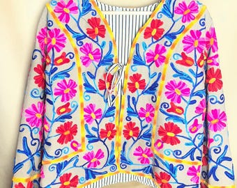 May include: A beige embroidered jacket with a floral design. The jacket features bright pink, red, and blue flowers with yellow accents. The interior lining has a black and white striped pattern. The jacket is hanging on a wooden hanger.