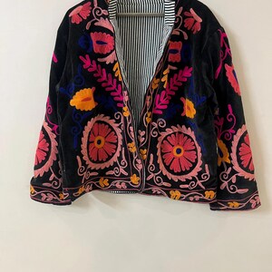 以下が含まれることがあります： ピンク、オレンジ、ブルーの鮮やかな花柄刺繍が施された黒いベルベットジャケット。 ジャケットはストライプの裏地とクラシックなオープンフロントデザインが特徴です。 刺繍は細かく、前面と袖を覆っています。