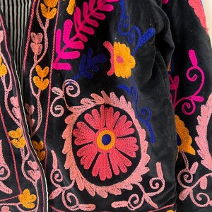 以下が含まれることがあります： 鮮やかな花の刺繍が施された黒いジャケットのクローズアップ。刺繍はピンク、イエロー、ブルーの糸を使用し、カラフルなデザインを作り出しています。ジャケットの内側の裏地は、白と黒のストライプ柄です。