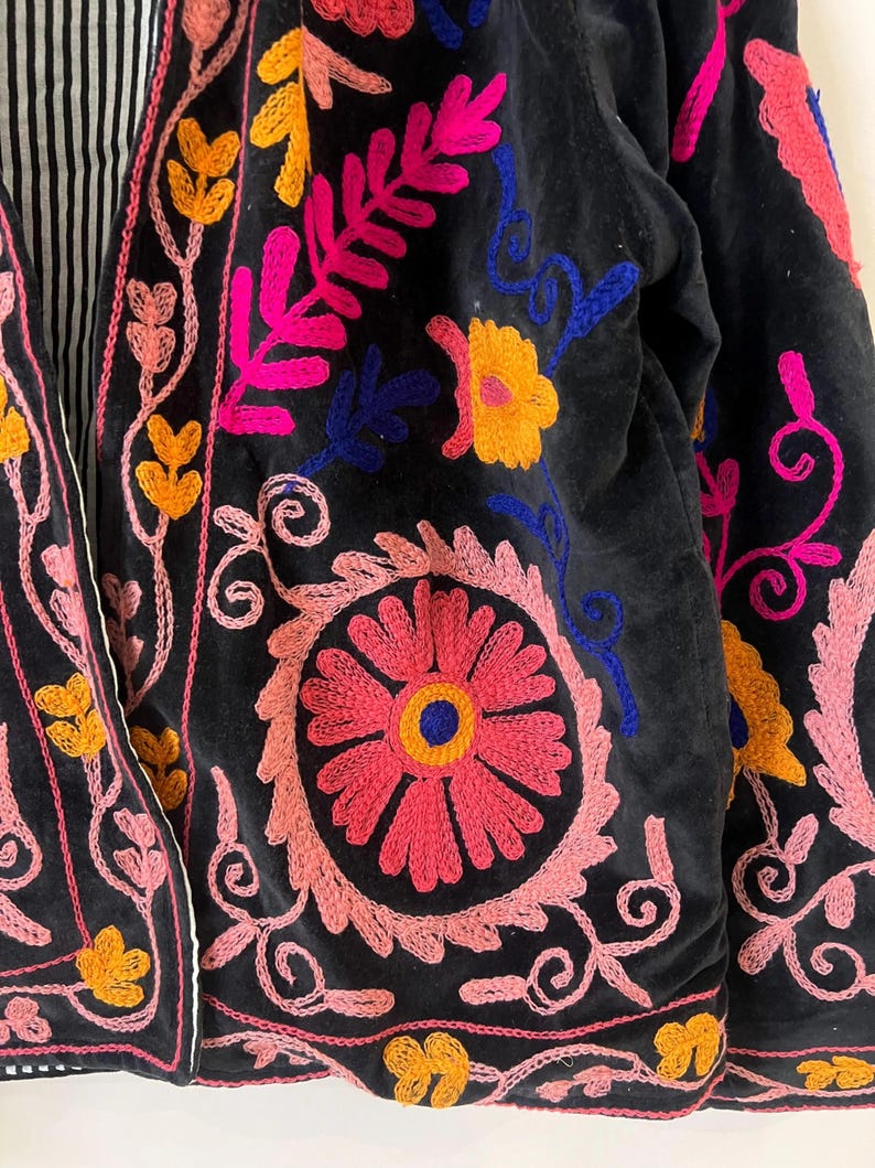 以下が含まれることがあります： 鮮やかな花の刺繍が施された黒いジャケットのクローズアップ。刺繍はピンク、イエロー、ブルーの糸を使用し、カラフルなデザインを作り出しています。ジャケットの内側の裏地は、白と黒のストライプ柄です。