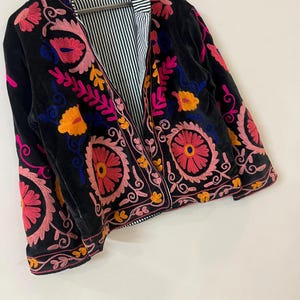 以下が含まれることがあります： ピンク、オレンジ、黄色、青の鮮やかな花柄刺繍が施された黒いベルベットジャケット。 ジャケットにはストライプの裏地が付いており、木製のハンガーに掛けられています。 刺繍のデザインは複雑で詳細で、ボヘミアンな美学を作り出しています。