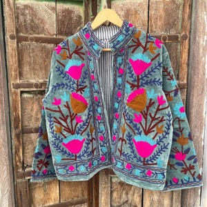 Puede incluir: Chaqueta de terciopelo verde azulado con bordados florales vibrantes en rosa, azul y marrón. La chaqueta tiene un cuello mandarín y está forrada con una tela a rayas. El bordado crea un contraste llamativo sobre el fondo verde azulado.