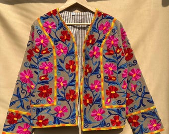 May include: A tan jacket with vibrant floral embroidery. The jacket features a V-neck design and is adorned with pink, red, and yellow flowers, along with blue leaves. The interior lining has a striped pattern.