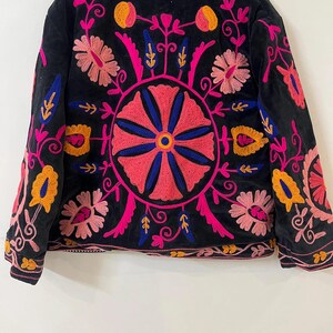 以下が含まれることがあります： ピンク、オレンジ、青色の鮮やかな花柄刺繍が施された黒いジャケット。背中には丸い花柄のデザイン、袖と襟には詳細な模様があります。刺繍は、暗い生地にカラフルなコントラストを加えています。