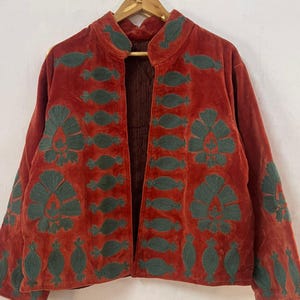 Könnte beinhalten: Rostfarbene Samtjacke mit dunkelgrünen, gestickten Blumen- und Blattmustern. Die offene Jacke hängt an einem Holzbügel vor weißem Hintergrund. Das Innere der Jacke ist ein dunklerer, gestreifter Stoff.