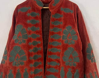May include: A rust-colored velvet jacket with dark green embroidered floral and leaf patterns. The jacket has a collar and is open in the front, revealing a striped lining. The sleeves are long and the jacket is hanging on a wooden hanger.