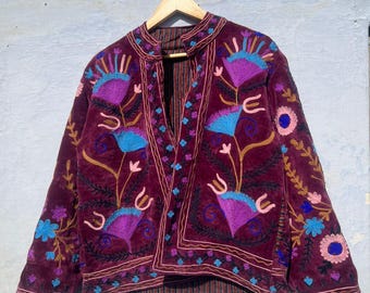 May include: A burgundy velvet jacket with intricate floral embroidery in shades of purple, blue, and gold. The jacket features a mandarin collar and is displayed on a wooden hanger. The embroidery creates a striking contrast against the dark fabric.