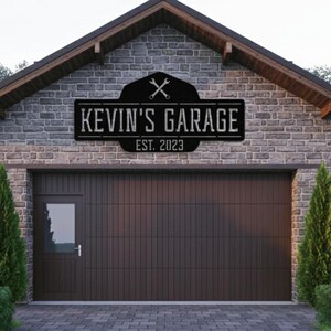Puede incluir: Un letrero de metal negro con el texto "KEVIN'S GARAGE EST. 2023" está montado en un edificio de ladrillo sobre una puerta de garaje marrón. El letrero presenta llaves cruzadas y un diseño de escudo. El garaje tiene una pequeña puerta a la izquierda.