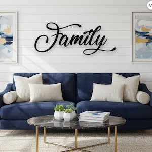 Puede incluir: Un letrero de pared negro con la palabra "Family" en cursiva está montado en una pared blanca de tablillas sobre un sofá azul marino con cojines color crema. Una mesa de centro redonda con tapa de mármol y patas doradas está frente al sofá.