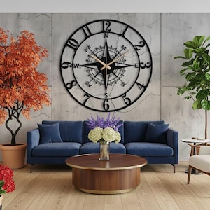 Könnte beinhalten: Eine große Wanduhr aus schwarzem Metall mit Kompassdesign ist an einer Betonwand befestigt. Die Uhr hat einen kreisförmigen Rahmen mit römischen Ziffern und Kompasspunkten. Der Raum verfügt über ein blaues Sofa, einen Couchtisch aus Holz und Topfpflanzen.