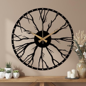 May include: A large, round, black metal wall clock with a tree branch design. The clock hands are gold. The clock is mounted on a beige wall. The clock is approximately 61 cm in diameter.