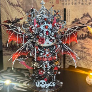 Puede incluir: Figurita detallada con una cara blanca y detalles rojos, adornada con una corona plateada, cuernos rojos y alas de murciélago. La figurita viste un atuendo negro y rojo con detalles de calaveras y rosas, y se asienta sobre una base con calaveras.