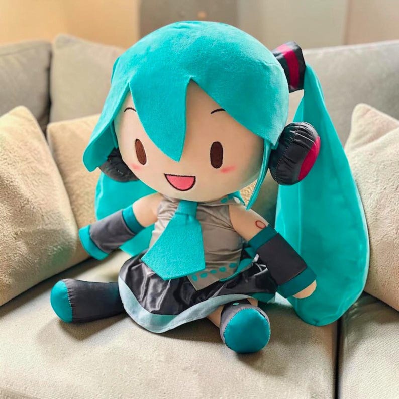 May include: A plush doll of a character with turquoise hair, a matching tie, and a gray and black outfit. The doll has large eyes, rosy cheeks, and is seated on a light-colored couch. The doll has black headphones with red accents.