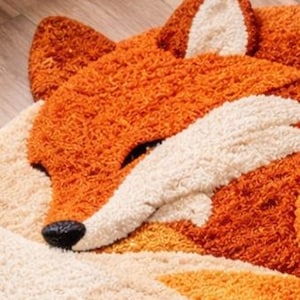 May include: A plush, orange and white fox-shaped rug. The rug features a detailed design with a black nose, white accents, and a textured, shaggy appearance. The fox is depicted in a sleeping pose, with its eyes closed.