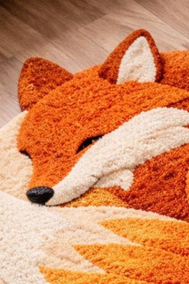 May include: A plush, orange and white fox-shaped rug. The rug features a detailed design with a black nose, white accents, and a textured, shaggy appearance. The fox is depicted in a sleeping pose, with its eyes closed.