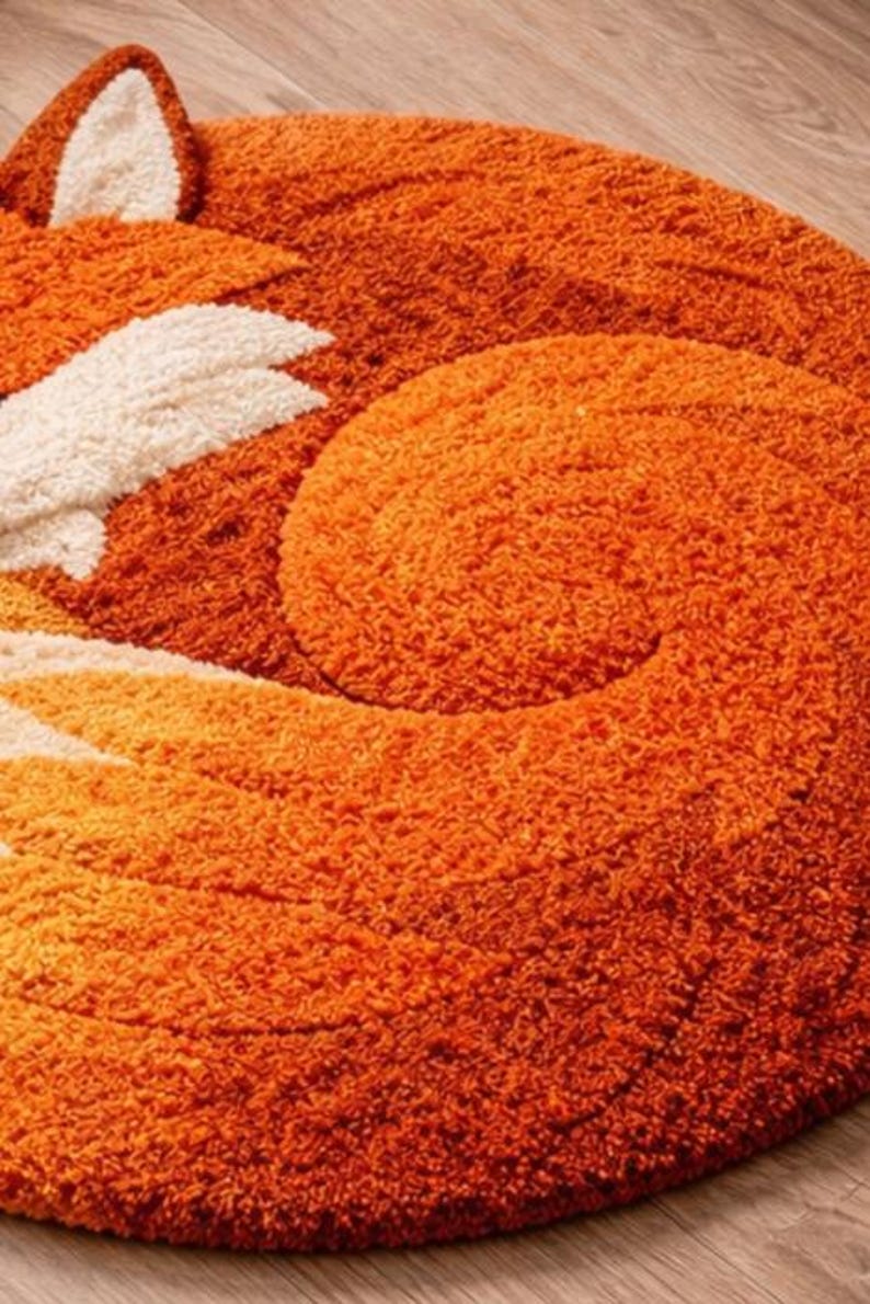 May include: A round, orange fox-shaped rug with a fluffy texture. The rug features a fox's head, ears, and curled tail in shades of orange and cream. This decorative rug is designed for a child's room or nursery.