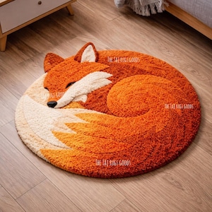 May include: A round, orange and cream-colored rug shaped like a sleeping fox. The rug has a textured, plush surface and features the fox's head, body, and tail in a curled position. The text "THE TAJ RUGS GOODS" is printed on the rug.