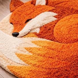 May include: A round, orange fox rug with a white and cream-colored face and tail. The rug is made of a soft, textured material and features a sleeping fox design. The fox's eyes are closed, and its body is curled up.