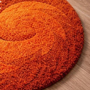 May include: A round, orange shag rug with a spiral design. The rug is made of a thick, textured material and sits on a wooden floor. The rug's color is a vibrant, warm orange.