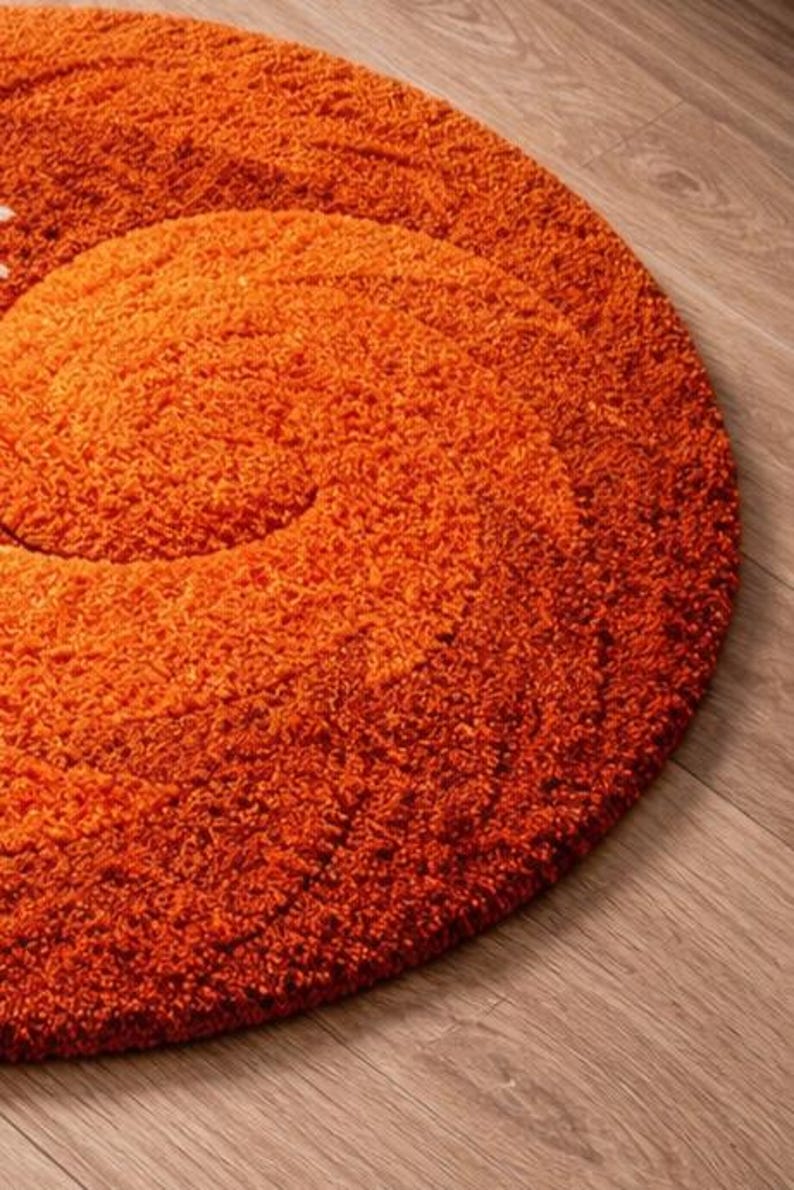May include: A round, orange shag rug with a spiral design. The rug is made of a thick, textured material and sits on a wooden floor. The rug's color is a vibrant, warm orange.