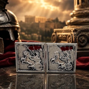 May include: A pair of square silver cufflinks with a detailed relief of a chariot pulled by two horses, a driver, and a red flag. The background features a blurred image of a classical building and a helmet.