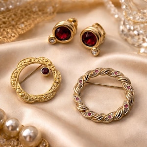 May include: An assortment of gold-toned jewelry, including a pair of earrings with large red stones and small clear stones, and two brooches. One brooch is a circle with a red stone, and the other is a braided circle with red and clear stones.