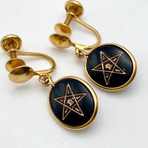 May include: A pair of gold-tone screw-back earrings featuring oval-shaped black cabochons. Each black oval is centered with a gold-tone five-pointed star design that contains intricate etched detailing and a small symbol in the center.