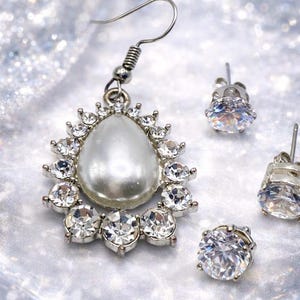 May include: A silver-toned earring with a pear-shaped pearl center, surrounded by clear crystals. Three additional stud earrings with clear crystals are also present, creating a coordinated jewelry set. The jewelry sparkles against a shimmering background.