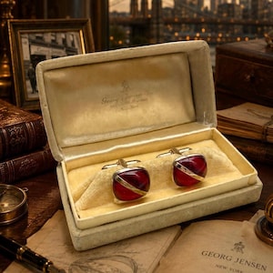 May include: A pair of red and silver cufflinks in an open, cream-colored box. The cufflinks have a rectangular shape with a red center and a silver diagonal stripe. The box is lined with yellow fabric. The box is on a table with a vintage aesthetic.