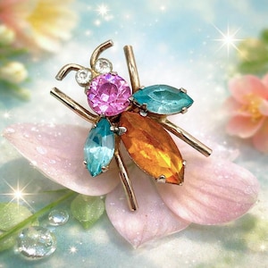 May include: A vintage insect brooch with a gold-tone metal frame. The brooch features a large pink center stone, teal and orange marquise-cut stones, and clear accent stones. The brooch is set on a pink petal.